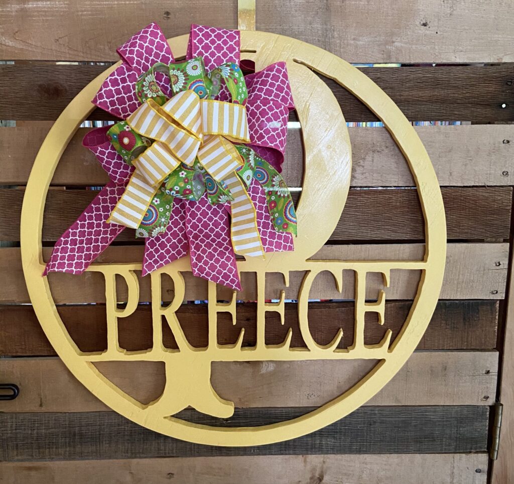 Round yellow, with the initial P and name Preece routed out of the center with a pink, green, and yellow bow attached to the sign.